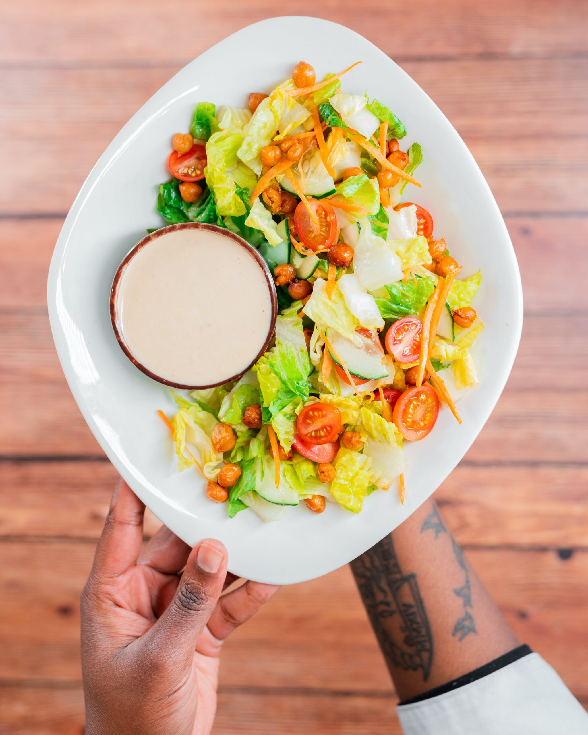 This photo shows a person holding a Caesar salad comprised of romaine lettuce, grape tomatoes, chickpeas, carrots, and cucumbers. With a small dish filled with caesar dressing on the side.